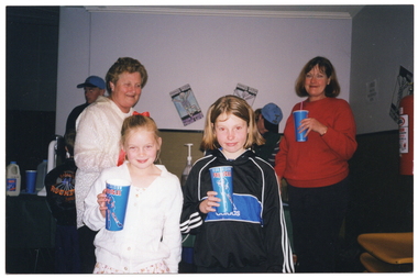 COL photo of two girls and two women standing indoors. The two girls and one of the women are each holding a disposable milkshake container in one hand. There two other people in the background, (partially obscured) standing behind a table with an electric milkshake maker appliance, bottles of milk and flavoured syrup. On the wall, there are two posters with a skeleton graphic and the text, "NATIONAL HEALTHY BONES WEEK YOUR BONES YOUR LIFE".