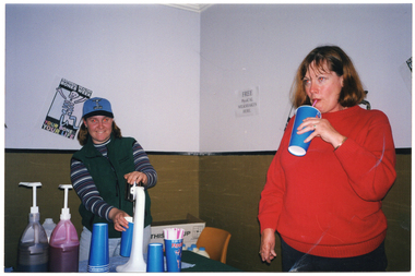 COL photo of two women standing behind a table in the corner of a hall. One woman is making a milkshake using a milkshake maker, while the other is drinking from a large blue disposable cup. On the wall behind them is a Healthy Bones, Healthy Body poster and a sign that says "FREE PhysiCAL MILKSHAKES HERE."