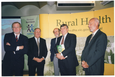 COL photo of five men posing for a photo in front of a large backdrop and banner. One of the men is holding a large booklet. On the left hand side of the backdrop is a pale green vertical freestanding banner. It is titled, "Human Services", with the subtitle 'People First' and a logo of two stick-type figures. The adjoining the backdrop is yellow and contains some large black and white Photographs. It is titled, 'Rural Health', with a Victorian government website address below the title. The website address (partially obscured) is "www.dhs.vic.gov. ...al".