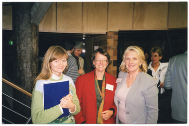 COL photo of three women standing close to one another in the lobby of a building. They are all wearing name tags on their jumpers or jackets. One of them is holding a blue folder close to her chest. There is a set of stairs to the left of the frame. In the background, there are several other people also in the lobby.