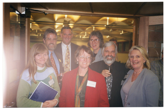 COL photo of a small group of people standing close together in two rows in the lobby of a building. There are four people in the front row, and three people in the back row. There are one or two other people (partially obscured) standing behind or beside them. In the background, inside the main hall of the venue, an ornate but modern decoratively lit ceiling is evident through a wide double doorway.