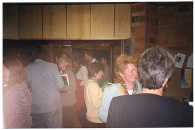 COL photo of a group of people standing in the lobby of a building. Some of them are chatting to one another. A few people are walking through a doorway from the main hall into the lobby.