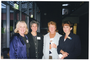 COL photo of four women posing for a photo outside a building at night. The building has tall floor to ceiling glass wall panels, and there is an "EXIT" sign above a double glass door.