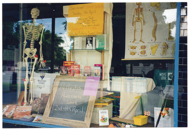 COL photo of a shop window display. Among the items on display are a plastic skeleton on a vertical stand, books, brochures, information sheets, posters, and a VicHealth Masters Games promotional t-shirt. Affixed to the top of a bookshelf is a yellow cardboard sign, containing the text "Join our Joint Walk Tuesday 11th April 9:30am Lions' Park (near Water Tower) 10:30am Morning Tea (B.Y.O. snack & drink) Everyone Welcome Rochester Arthritis Support Group". Leaning against the bottom of the bookcase is a small wooden-framed blackboard, with the text "Arthritis Awareness Week 2nd - 8th April" written in chalk.
