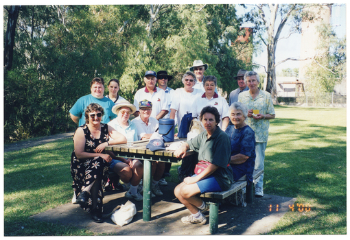 COL photo of a group of people gathered around a wooden picnic table in a park. Most of the people are standing around the picnic table, while some people are sitting on the two benches on either side. There are a few items on the table, such as a water bottle, some backpacks, a cap, and a small drink cooler box (known in Australian vernacular as an 'esky', or a 'chilly bin' in New Zealand vernacular). In the background, there are some large trees, and a large tank beyond a cyclone fence.