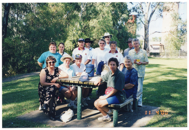 COL photo of a group of people gathered around a wooden picnic table in a park. Most of the people are standing around the picnic table, while some people are sitting on the two benches on either side. There are a few items on the table, such as a water bottle, some backpacks, a cap, and a small drink cooler box (known in Australian vernacular as an 'esky', or a 'chilly bin' in New Zealand vernacular). In the background, there are some large trees, and a large tank beyond a cyclone fence.