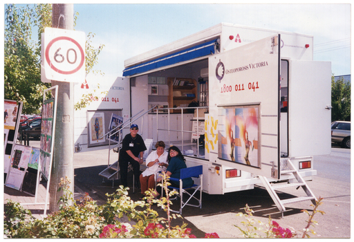 COL photo of a large van parked in a street. The bus is set up and its wide side doors are open to reveal the contents inside. Three people are in attendance: two are seated on deck chairs while the other is standing, leaning back onto the steps leading up to the inside of the van. There are display boards set up on the footpath. The text on one of the van's doors is "Arthritis Victoria 1800 011 041", and on the other door, it is "Osteoporosis Victoria 1800 011 041". Below the text on each door are some large posters.