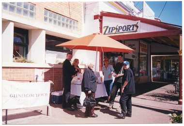 COL photo of a small group of people gathered around a table beneath an outdoor umbrella in a shopping strip. There is a table with various items on it, such as a blender, a bottle of orange juice, and some information inside a clear plastic frame. There is an extension lead connecting the blender to an electric power outlet through an open window, and a large esky is evident behind the table, against the exterior wall of the building. The shop next door has a sign on the front which says, "EASTERN ELECTRICS" and another which says "SALES AND SERVICE". On the side of the shop's veranda roof is a sign which says, "ZIPSPORTS".