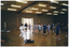 COL photo of a group of school children and a woman participating in a martial arts lesson or workshop. They are dressed in their school uniforms (wearing socks, but with their shoes off), while the instructor is wearing a white martial arts uniform with a black belt and is barefoot. They are in a large hall or gymnasium. In the background, there is a skeleton on a vertical stand.