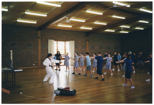 COL photo of a group of school children and a woman participating in a martial arts lesson or workshop. They are dressed in their school uniforms (wearing socks, but with their shoes off), while the instructor is wearing a white martial arts uniform with a black belt and is barefoot. They are in a large hall or gymnasium. In the background, there is a skeleton on a vertical stand.