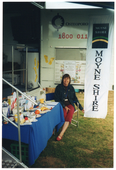 COL photo of a woman sitting on a deck chair in front of a large van. The van has a large door open on the side, displaying the text, "Osteoporosis Victoria 1800 011 041" (partially obscured). Below the text, there is a display board with some posters on it. Hanging from the edge of the van door is a long vertical banner with the text and logo for the Moyne Shire. In front of the side of the van (beside the woman), there is a rectangular table covered with a blue tablecloth. On it, there is a range of healthy food items such as cereals, various types of milk, cheeses, yoghurt, a plastic ice cream container, some canned food, and some fresh fruit. In the foreground, to the left of the frame, there are some metal steps with a ballustrade.
