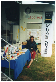 COL photo of a woman sitting on a deck chair in front of a large van. The van has a large door open on the side, displaying the text, "Osteoporosis Victoria 1800 011 041" (partially obscured). Below the text, there is a display board with some posters on it. Hanging from the edge of the van door is a long vertical banner with the text and logo for the Moyne Shire. In front of the side of the van (beside the woman), there is a rectangular table covered with a blue tablecloth. On it, there is a range of healthy food items such as cereals, various types of milk, cheeses, yoghurt, a plastic ice cream container, some canned food, and some fresh fruit. In the foreground, to the left of the frame, there are some metal steps with a ballustrade.