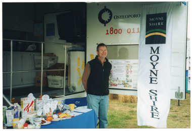 COL photo of a man standing next to a rectangular table and a deck chair, which have been set up in front of a large van. The van has a large door open on the side, displaying the text, "Osteoporosis Victoria 1800 011 041" (partially obscured). Below the text, there is a display board with some posters on it. Hanging from the edge of the van door is a long vertical banner with the text and logo for the Moyne Shire. The table is covered with a blue tablecloth. On it, there is a range of healthy food items such as cereals, various types of milk, cheeses, yoghurt, a plastic ice cream container, some canned food, and some fresh fruit. In the background, built-in cupboards, boxes and crates, and a television set affixed to the wall are evident inside the van.