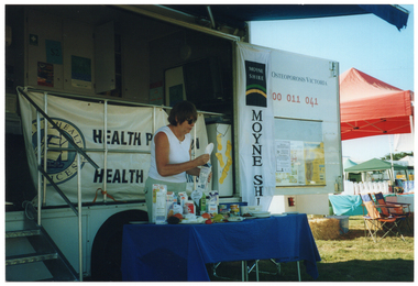 COL photo of a woman standing behind a rectangular table in front of a large van. On it, there is a range of healthy food items such as cereals, various types of milk, cheeses, yoghurt, a plastic ice cream container, some canned food, and some fresh fruit. The woman is affixing a display label to the top of a milk carton. The van has a large door open on the side, displaying the text, "Osteoporosis Victoria 1800 011 041" (partially obscured). Below the text, there is a display board with some posters on it. Hanging from the inside edge of the van door is a long vertical banner with the text and logo for the Moyne Shire. Behind the woman, there is a large "Moyne Health Services" banner attached to the ballustrade on the side of the van. In the foreground, to the left of the frame, there are some metal steps with a ballustrade. In the background, some market stall tents and marquees are evident, with tables and deck chairs underneath them.