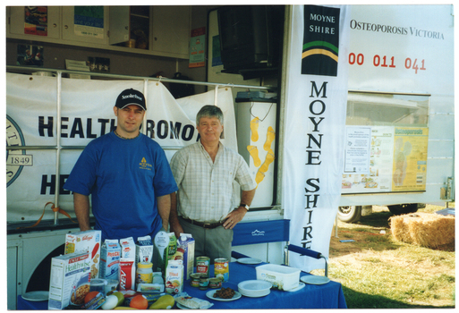 COL photo of two men standing behind a rectangular table in front of a large van. On it, there is a range of healthy food items such as cereals, various types of milk, cheeses, yoghurt, a plastic ice cream container, some canned food, and some fresh fruit. The van has a large door open on the side, displaying the text, "Osteoporosis Victoria 1800 011 041". Below the text, there is a display board with some posters on it. Hanging from the inside edge of the van door is a long vertical banner with the text and logo for the Moyne Shire. Behind the men, there is a large "Moyne Health Services" banner (partially obscured) attached to the ballustrade on the side of the van. In the background, there is a bale of hay on the grass, and two vehicle or trailer wheels are evident behind it.