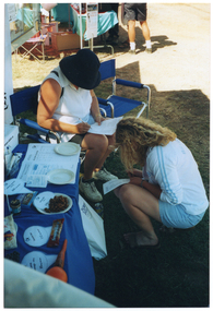 COL photo of two women outdoors, on a grassy area. One woman is sitting on one of two blue deck chairs, while the other woman is crouching down in front of her. They are both writing on some printed sheets of paper. Beside them, there is a table covered with a bright blue tablecloth, which has a variety of healthy food items, with labels, on it. In the background, there is someone standing next to another small table covered with a green cloth.