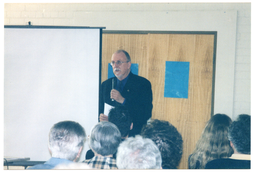 COL photo of a man standing beside a slide projector screen, speaking into a hand-held microphone. Behind him is a double door, with a blue poster or sign on each door. The audience, who have been Photographed from behind, are evident in the foreground.