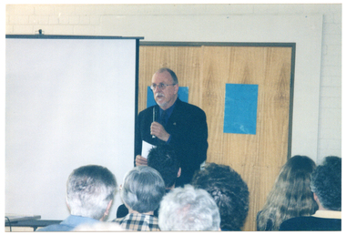 COL photo of a man standing beside a slide projector screen, speaking into a hand-held microphone. Behind him is a double door, with a blue poster or sign on each door. The audience, who have been Photographed from behind, are evident in the foreground.