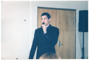 COL photo of a man, standing, speaking into a hand-held microphone. Behind him, there is a closed double door and, beside the doors, there is a speaker on a stand.