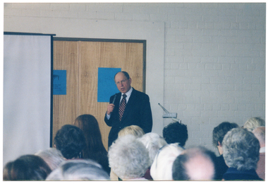COL photo of a man standing beside a slide projector screen, speaking into a hand-held microphone. Behind him is a double door, with a blue poster or sign on each door. The audience, who have been Photographed from behind, are evident in the foreground.