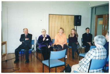 COL photo of a small group of people sitting in a large room. On one side, there are five people sitting on a row of five chairs. One of them is speaking into a hand-held microphone. Another person has his arms folded across his chest. Behind them, there is a double door and a speaker on a stand. On a perpendicular wall, there is a set of double glass doors with a wooden frame. In the foreground, there are two rows of chairs, with two people (one partially obscured) occupying chairs in the second row. They have been Photographed from behind, at a slight angle.