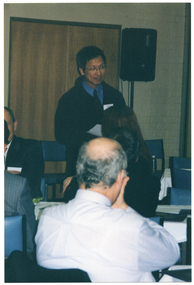COL photo of a small group of people in a room. Four of them are sitting in a row on one side. One of them is a woman who is speaking into a hand-held microphone. Behind them, there is a double door and a speaker on a stand. In the foreground, two people who are also seated, have been Photographed from behind.