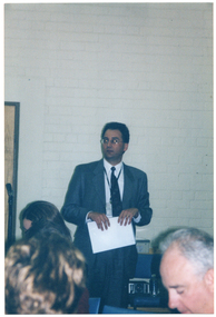 COL photo of a man standing in a room with several people, who are seated. He is holding a sheet of paper in his hand. Behind him, there is a double set of doors and a speaker on a stand.