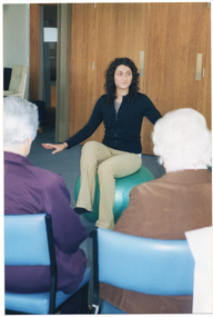 COL photo of a man standing in a room with several people, who are seated. He is holding a booklet or folder with both hands. Behind him, to the left side of the frame, there is a double set of doors and a microphone stand.