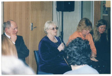 COL photo of a woman demonstrating some exercises to a group of people. She is sitting on a large exercise ball with her knees bent and one leg held slightly higher than the other, and her arms held out to the side. In the foreground, two people who are also seated, have been Photographed from behind.