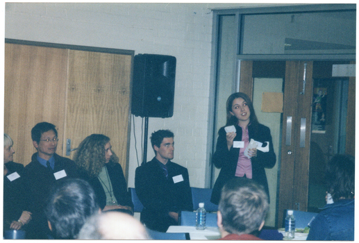 COL photo of a group of people inside a room. One woman, standing, is holding an object in each of her hands and has some sheets of paper tucked inside her arm. Beside her, there are four people sitting on a row of chairs. Behind them, there is a double door and a speaker on a stand. On a perpendicular wall, there is a set of double glass doors with a wooden frame. In the foreground, there are several people who are also seated and have been Photographed from behind. There is a table covered in a white tablecloth between the two groups, with two water bottles, a polystyrene cup, and a plate of food on top.