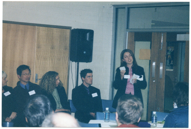 COL photo of a group of people inside a room. One woman, standing, is holding an object in each of her hands and has some sheets of paper tucked inside her arm. Beside her, there are four people sitting on a row of chairs. Behind them, there is a double door and a speaker on a stand. On a perpendicular wall, there is a set of double glass doors with a wooden frame. In the foreground, there are several people who are also seated and have been Photographed from behind. There is a table covered in a white tablecloth between the two groups, with two water bottles, a polystyrene cup, and a plate of food on top.