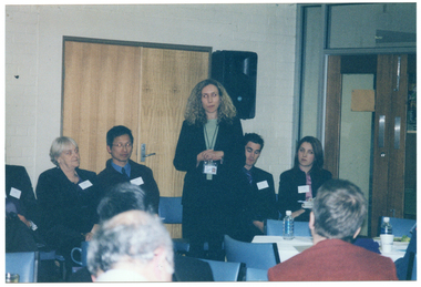 COL photo of a group of people inside a room. One woman is standing, with several people sitting on chairs on either side of her. Behind them, there is a double door and a speaker on a stand. On a perpendicular wall, there is a set of double glass doors with a wooden frame. In the foreground, there are a few people who are also seated and have been Photographed from behind. There is a table covered in a white tablecloth between the two groups, with two water bottles, a polystyrene cup, and a plate of food on top.