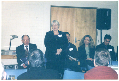 COL photo of a group of people inside a room. One woman is standing, with people sitting on chairs on either side of her. Behind them, there is a double door and a speaker on a stand. On a trolley near the doors, there is an overhead slide projector. In the foreground, there are a few people who are also seated and have been Photographed from behind. There is a table covered in a white tablecloth between the two groups, with a water bottle on top.