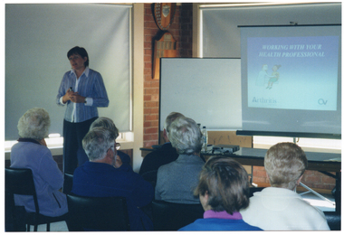 COL photo of a woman standing in front of a group of people who are seated. She is standing next to a window with the blinds drawn. Behind her, there is a freestanding whiteboard and a slide projector screen, with another large window with the blinds drawn behind those. The slide being projected onto the screen is titled, "WORKING WITH YOUR HEALTH PROFESSIONAL", beneath which there is a cartoon of a doctor attending to a patient. In the bottom left corner of the slide, there is the Arthritis Victoria text (incorporating the logo in the letter 'A'), and in the bottom right corner of the slide, there is Osteoporosis Victoria's 'OV' logo. In the foreground, several people, who are seated, have been Photographed from behind.
