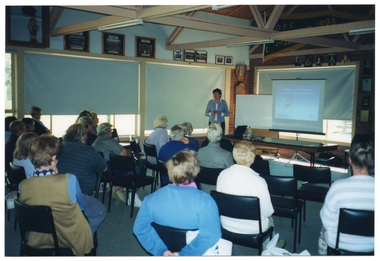COL wide frame photo of a woman standing in front of a large group of people who are seated. The room has several large windows along one wall, and another large window on the back wall. The blinds are drawn on three of the windows. There is a freestanding whiteboard and a slide projector screen in front of the back window, behind a long table. The slide being projected onto the screen is titled, "WORKING WITH YOUR HEALTH PROFESSIONAL", beneath which there is a cartoon of a doctor attending to a patient. In the bottom left corner of the slide, there is the Arthritis Victoria text (incorporating the logo in the letter 'A'), and in the bottom right corner of the slide, there is Osteoporosis Victoria's 'OV' logo. In the foreground, several people, who are seated, have been Photographed from behind.  High on the wall, above the windows, there are several framed photos and a large banner depicting the letters "O.F.C" and the year 1993. In the foreground, a large group of people, who are all seated in rows of chairs, have been Photographed from behind. The photo has been taken from a 45 degree angle.