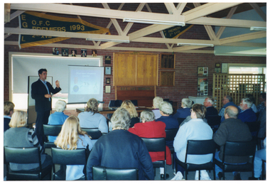 COL wide frame photo of a man standing in front of a group of people who are seated. He is standing in front of a window with the blinds drawn. Behind him, there is a freestanding whiteboard. The slide being projected onto the whiteboard is titled, "What causes Rheumatoid Arthritis?" and contains some diagrams and drawings. Next to the whiteboard, there is a slide projector screen and a large wooden board behind a long rectangular table. The wooden board contains the text, "O.F.C. LIFE MEMBERS" below. On the brick wall, there are two smaller wooden leader boards. High on the wall, above the windows, there are several framed photos and a large banner depicting the letters "O.F.C" and the year 1993. In the foreground, a large group of people, who are all seated in rows of chairs, have been Photographed from behind. The photo has been taken from a slight angle.