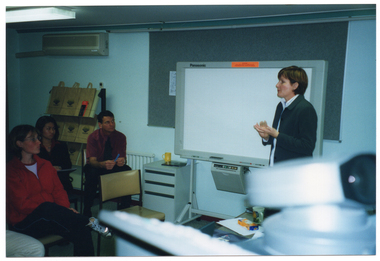 COL photo of a woman standing in front of a small group of people who are seated. She is standing in front of an electronic whiteboard, which is against the wall. Behind the whiteboard, there is a large grey display board. To the left, there is a small filing cabinet, a wall radiator, and a display stand with some branded tote bags on the shelves. Above the stand, there is an air conditioning unit. To the left of the frame, there are a few people sitting on chairs. They are looking at the woman standing.