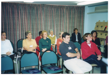 COL wide frame photo of a group of people sitting on three rows of chairs. There is a large booklet on the seat of a few unoccupied chairs in the front row. In the background, there is a floor-to-ceiling curtain along one wall. To the right of the frame, there is a stand with some branded tote bags on the shelves. There is an airconditioner on the wall above the stand. The photo has been taken from a slight angle.