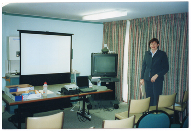COL wide frame photo of a woman standing in a room. There is a is a projector screen behind a long table and, beside it, a television set on a stand with castors. On the table, there is a cardboard box and its lid with some miscellaneous items placed inside both, a plastic water bottle, some slide projector equipment, a laptop computer, and a computer keyboard. In the background (behind the projector screen), there is a cupboard with a tap and sink, and an electric hot water urn above. Behind the woman, there are some floor to ceiling curtains along the wall. In the foreground, there are several empty chairs arranged in rows.