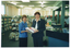 COL wide frame photo of two women standing in a library. Together, they are holding a stack of books. There is a customer service counter beside them, with various items on top, including leaflets on a stand and on the counter surface, a stack of video cassettes in their cases, and a few books. In the background, multiple bookshelves line the walls, with windows above them. In the middle of the room, there is a circular rotating stand with some bookshelves behind it - both containing various information resources.