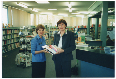 COL wide frame photo of two women standing in a library. Together, they are holding a stack of books. There is a customer service counter beside them, with various items on top, including leaflets on a stand and on the counter surface, a stack of video cassettes in their cases, and a few books. In the background, multiple bookshelves line the walls, with windows above them. In the middle of the room, there is a circular rotating stand with some bookshelves behind it - both containing various information resources.