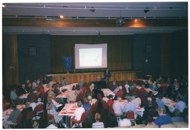 COL wide frame photo of a man speaking at a lectern in a large hall. Behind him, there is a stage with a large projector screen on a stand. A slide image titled, "What causes osteoarthritis?", with a diagram of a bone and joint, is being projected onto the screen. Beside the screen, there is a freestanding blue and gold banner on a stand. In the foreground, numerous people are seated at long rectangular tables. There are sheets of orange paper on the table at most place settings. There is a mirror ball hanging from the centre of the ceiling. This photo has been taken from the left, at a slight angle.