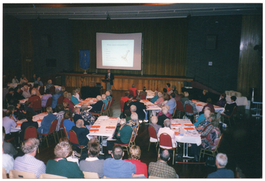 COL wide frame photo of a man speaking at a lectern in a large hall. Behind him, there is a stage with a large projector screen on a stand. A slide image titled, "What causes osteoarthritis?", with a diagram of a bone and joint, is being projected onto the screen. Beside the screen, there is a freestanding blue and gold banner on a stand. Numerous people are seated at long rectangular tables in front of the stage and lectern. There are sheets of orange paper on the table at most place settings. In the foreground, there are several people sitting on rows of chairs.  There is a mirror ball hanging from the centre of the ceiling. This photo has been taken from the right, at a slight angle.