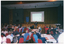 COL wide frame photo of a man speaking at a lectern in a large hall. Behind him, there is a stage with a large projector screen on a stand. A slide image, titled "How can I manage my arthritis?", with two illustrations of people exercising and walking, is being projected onto the screen. Beside the screen, there is a freestanding blue and gold banner on a stand. In the foreground, numerous people are seated at long rectangular tables. There are sheets of orange paper on the table at most place settings. This photo has been taken from the right, at a slight angle.