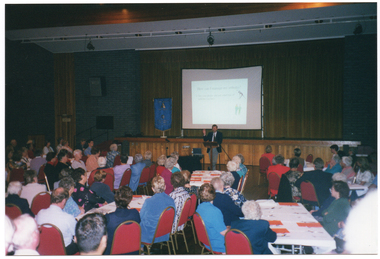 COL wide frame photo of a man speaking at a lectern in a large hall. Behind him, there is a stage with a large projector screen on a stand. A slide image, titled "How can I manage my arthritis?", with two illustrations of people exercising and walking, is being projected onto the screen. Beside the screen, there is a freestanding blue and gold banner on a stand. In the foreground, numerous people are seated at long rectangular tables. There are sheets of orange paper on the table at most place settings. This photo has been taken from the right, at a slight angle.