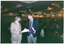 COL photo of two men standing next to one another, chatting. One of the men is holding some papers, while the other is holding a blue printed booklet or program. In the background, numerous people are seated at tables in a large dark room. Several downlights in the ceiling are evident.