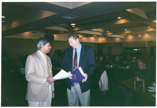 COL photo of two men standing next to one another, chatting. One of the men is holding some papers, while the other is holding a blue printed booklet or program. In the background, numerous people are seated at tables in a large dark room. Several downlights in the ceiling are evident.