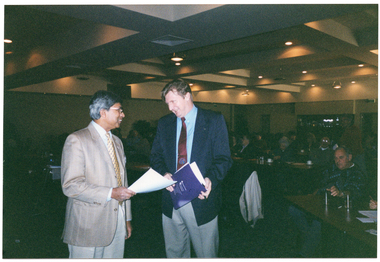 COL photo of two men standing next to one another, chatting. One of the men is holding some papers, while the other is holding a blue printed booklet or program. In the background, numerous people are seated at tables in a large dark room. Several downlights in the ceiling are evident.