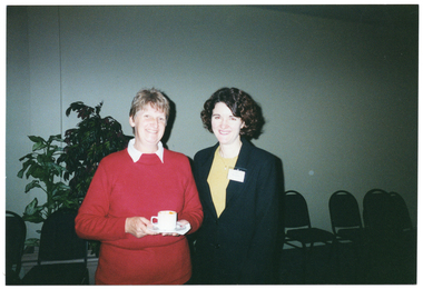 COL photo of two women standing next to one another, smiling. One of the women is holding a cup and saucer in her hands. Behind them, there is a row of chairs against the wall, and a large potted plant.