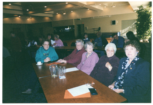 COL photo of numerous people seated at long rectangular tables in a large room. In the foreground, four women are seated at one of the tables. On top of the table, there are a jug and some glasses, a cup and saucer, some orange and some white papers, a pen, and some spectacles with their case.